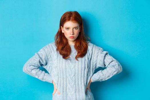 Redhead girl staring angry and displeased at camera, frowning upset, standing against blue background.