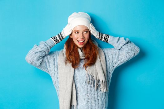Winter and holidays concept. Happy redhead girl in beanie hat, scarf and gloves looking right and smiling, standing against blue background.