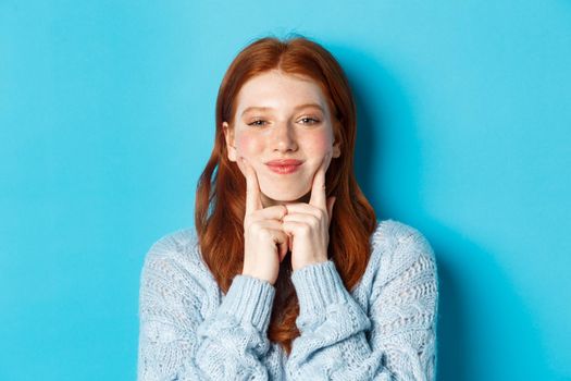 Close-up of cute and silly girl with red hair poking cheeks, showing dimples and smiling, standing against blue background.