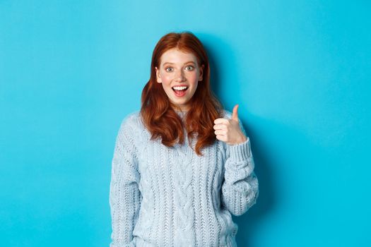 Impressed redhead girl showing thumb up and staring amazed at camera, praising good work, standing amazed against blue background.