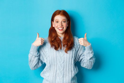 Cheerful teen girl with red hair, showing thumbs up in approval, like and praise gesture, standing over blue background.