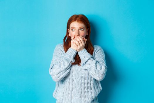 Shocked and anxious redhead girl staring left scared, covering mouth and gasping, standing over blue background in sweater.