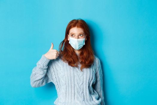 Winter, covid-19 and social distancing concept. Satisfied young redhead woman in face mask showing alright, thumb up gesture and looking left at promo, blue background.