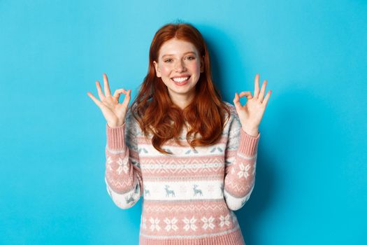 Cute redhead girl in sweater showing okay signs, smiling pleased, approve and like, praise excellent choice, standing over blue background.