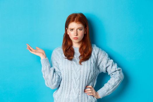 Skeptical teenage girl looking unamused, raising hand in so what gesture, staring at something with careless face, standing over blue background.