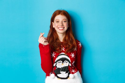 Winter holidays and Christmas Eve concept. Cute smiling redhead girl in xmas sweater, showing heart sign and wishing happy New Year, standing over blue background.