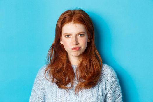 Disappointed teenage girl with red hair, frowning and smirking displeased, looking judgemental, standing against blue background.