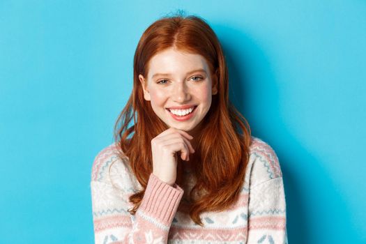 Close-up of beautiful redhead girl smiling, having conversation and staring at camera interested, standing over blue background.