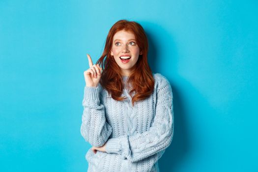 Thoughtful redhead girl having an idea, raising finger and smiling pleased with good plan, standing over blue background.