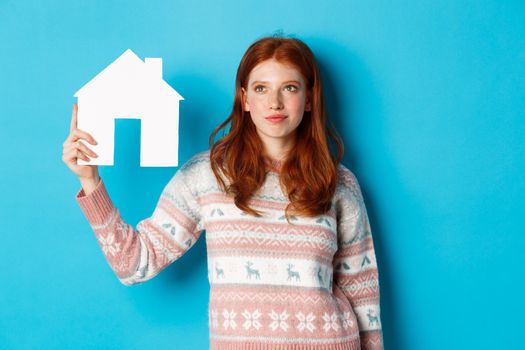 Real estate concept. Image of cute redhead girl looking curious at paper house model, thinking of buying property, looking up at copy space, blue background.