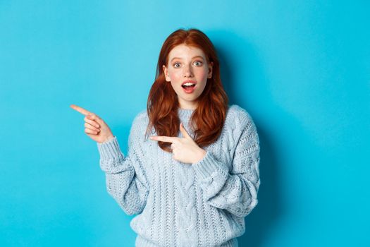 Amazed redhead girl in sweater, pointing fingers left at logo and staring excited at camera, standing against blue background.