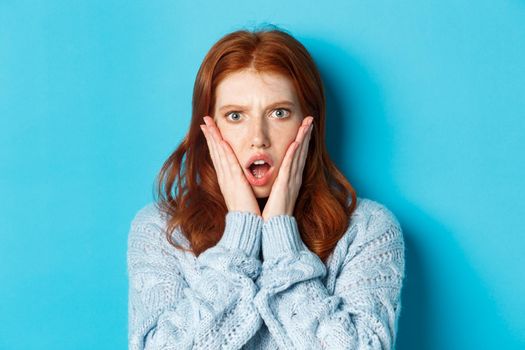 Close-up of shocked redhead girl staring at something displeasing, holding hands on face and gasping, standing over blue background.