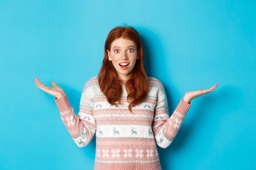 Image of excited redhead girl react to good news, looking surprised, spread hands sideways and smiling, standing in winter sweater against blue background.
