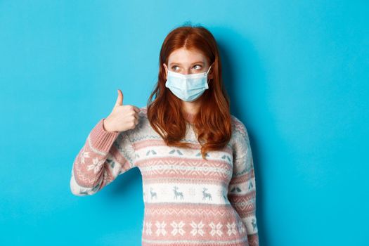 Winter, coronavirus and social distancing concept. Pretty redhead girl in face mask and sweater, looking at upper left corner promo and showing thumb up, praising good product.