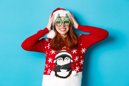 Happy holidays and Christmas concept. Funny redhead teen girl celebrating New Year, wearing santa hat and party glasses, standing against blue background.