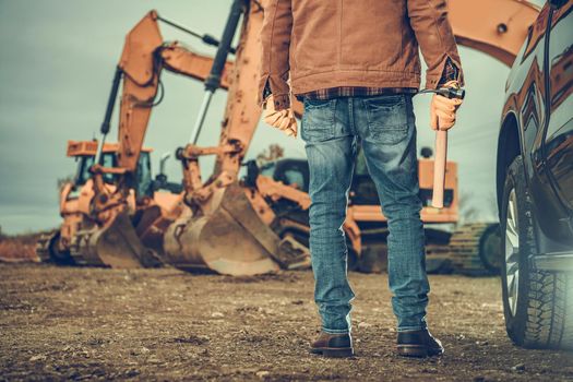 Professional Caucasian Contractor Worker in His 40s and a Construction Site with Excavator Machines and a Pickup Truck. Ground Works Industry Theme.