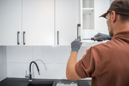 Caucasian Professional Cabinetmaker in His 40s Finishing Residential Kitchen Cabinets Installation. White Modern Kitchen Furniture.