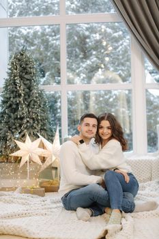 Handsome boy with his beautiful girlfriend hug each other at home at Christmas eve. Fit tree and snow outsif. Christmas mood.