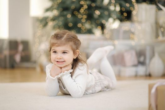 Adorable little girl lying on her front nearby fir tree and smile. Christmas mood at home with lot of presents.
