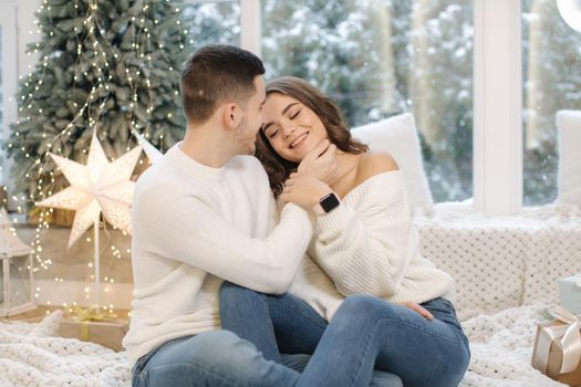 Handsome boy with his beautiful girlfriend hug each other at home at Christmas eve. Fit tree and snow outsif. Christmas mood.