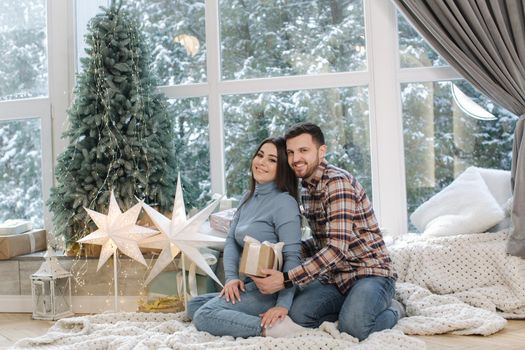 Attractive young woman with her beloved husband hold Christmas present. Female sitting in front of big window and near the fir tree. Snow outside.