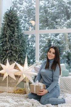Attractive young woman in beautiful blue denim style hold Christmas present. Female sitting in front of big window and near the fir tree. Snow outside.