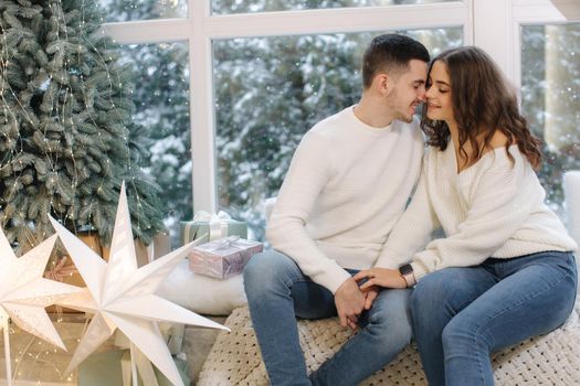 Beautiful young couple sitting in front of big window and fir tree. It's snow outside.