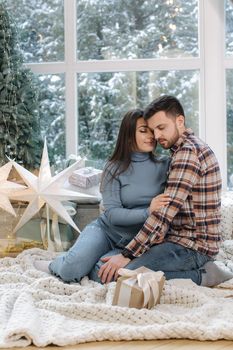 Young and beautiful couple of man and woman sitting in front of big windor, it's snow outside. Happy family at Christmas time.