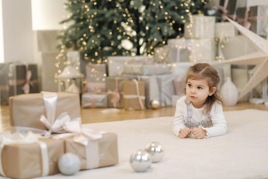 Adorable little girl lying on her front nearby fir tree and smile. Christmas mood at home with lot of presents.