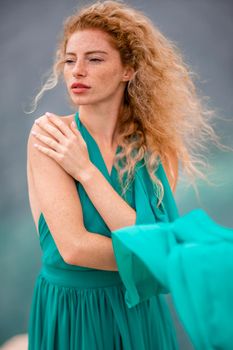Outdoor portrait of a young beautiful natural redhead girl with freckles, long curly hair, in an emerald dress, posing against the background of the sea