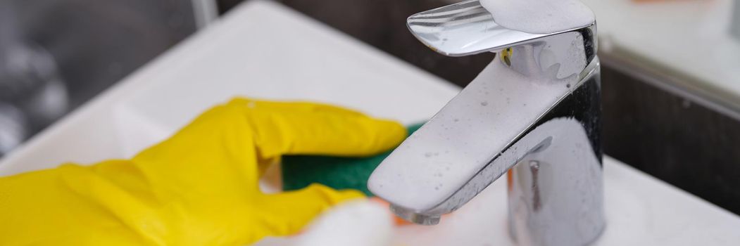 Person in yellow gloves washes sink with washcloth and sprays cleaning foam on faucet. Bathroom ceramics cleanliness and care concept