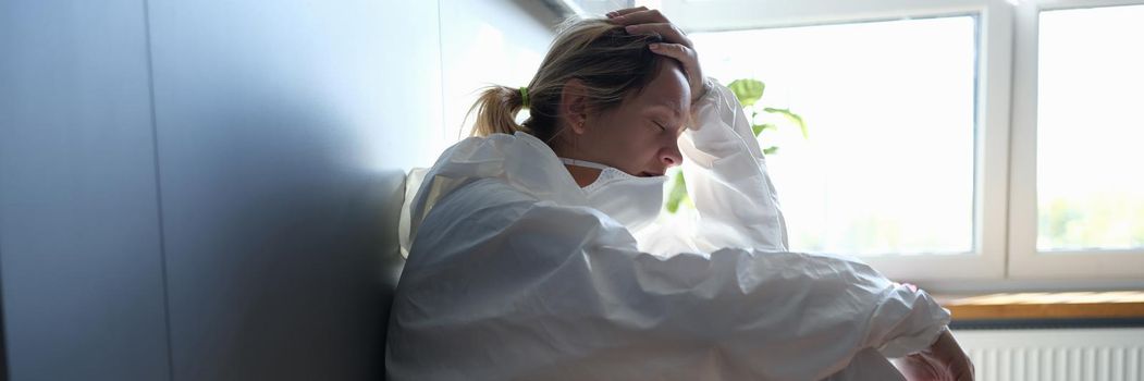 Tired female doctor in medical protective suit and mask in hands after work in clinic. Harsh working conditions of doctors in coronavirus pandemic concept