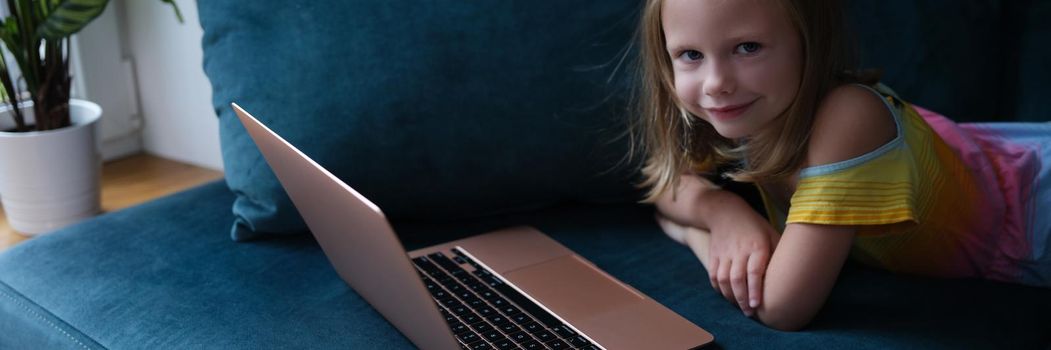 Little smiling girl lies on couch with laptop. Baby apps digital technology concept