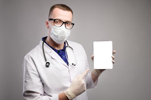 A young Caucasian doctor in a mask in a white coat and glasses looks at the camera and shows at an electronic tablet with a white cut-out screen. Studio portrait on gray background.