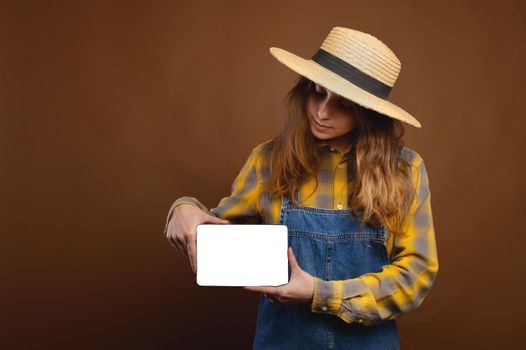 Studio portrait. Long-haired Caucasian attractive young woman in country clothes, denim overalls and shirt holds an electronic tablet with a cut-out screen