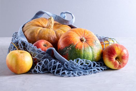 Close-up fresh ripe pumpkins and apples in environmentally friendly reusable mesh bag made from recycled materials on gray background. Healthy food concept, vegetarianism, no plastic. Selective focus.