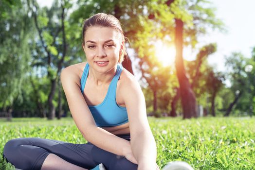 Beautiful smiling girl stretching before fitness training session in park at sunrise. Young woman in activewear sitting on green grass at summer day. Morning workout and healthy lifestyle.