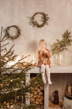 A girl is playing on Christmas Eve in a beautiful house decorated for the New Year holidays. Scandinavian-style interior with live fir trees and a wooden staircase.