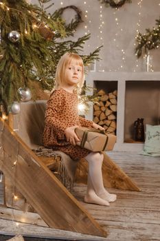 A little blonde girl is sitting on a wooden staircase in a Scandinavian interior decorated in a New Year's style. A child holds a Christmas gift in a craft package.