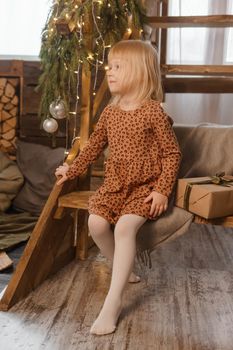 A little blonde girl is sitting on a wooden staircase in a Scandinavian interior decorated in a New Year's style. A child holds a Christmas gift in a craft package.