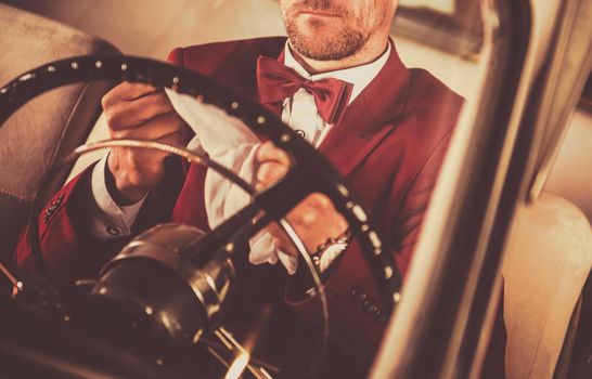 Elegant Men Wearing Bow Tie and Burgundy Siut Behind Classic Car Steering Wheel. 