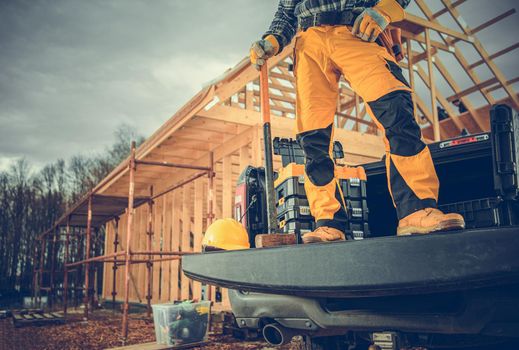 Contractor Worker and the Construction Site. Men with Large Hammer Staying on a Pickup Cargo Bed in Front of Wooden House Construction.