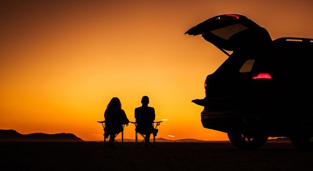 Couple Enjoying Their Life Watching Scenic Sunset on the Middle of a Desert Seating on Camping Chairs Next to Their Sport Utility Vehicle. Southern California USA