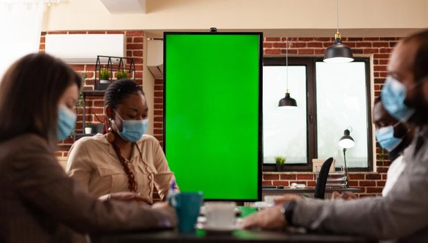 Multiethnic teamwork with medical protective face mask against coronavirus working at business strategy in startup company meeting office. Mock up green screen chroma key monitor with isolated display