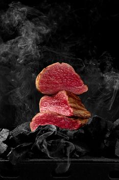 Sliced juicy rare veal tenderloin stacked on coals on bbq grill on black background in light white smoke