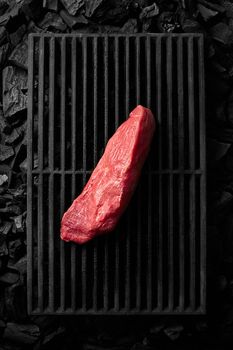 Top view of piece of lean pork tenderloin lying on black cast iron grill grate over cold coals ready for cooking