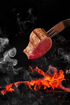 Slice of tender veal fillet cooking on fork over burning charcoals on grill against black background in light white smoke and soaring sparks