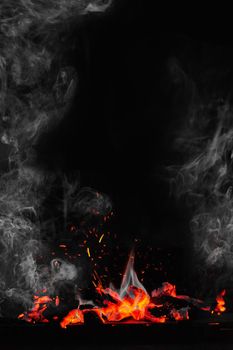 Fire flames and glowing red coals of burnt wood on grill on black background with soaring bright sparks and light white smoke. Vertical image with copyspace