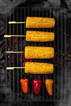 Appetizing ears of corn on wooden skewers and multicolored bell peppers cooking on black cast iron barbecue grate over smouldering coals with soaring sparks and white smoke