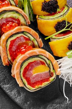 Macro shot of two types of rolls with salmon, tuna, mango and avocado garnished with red caviar, tobiko, grated daikon and greens served on black slate board. Authentic Japanese cuisine